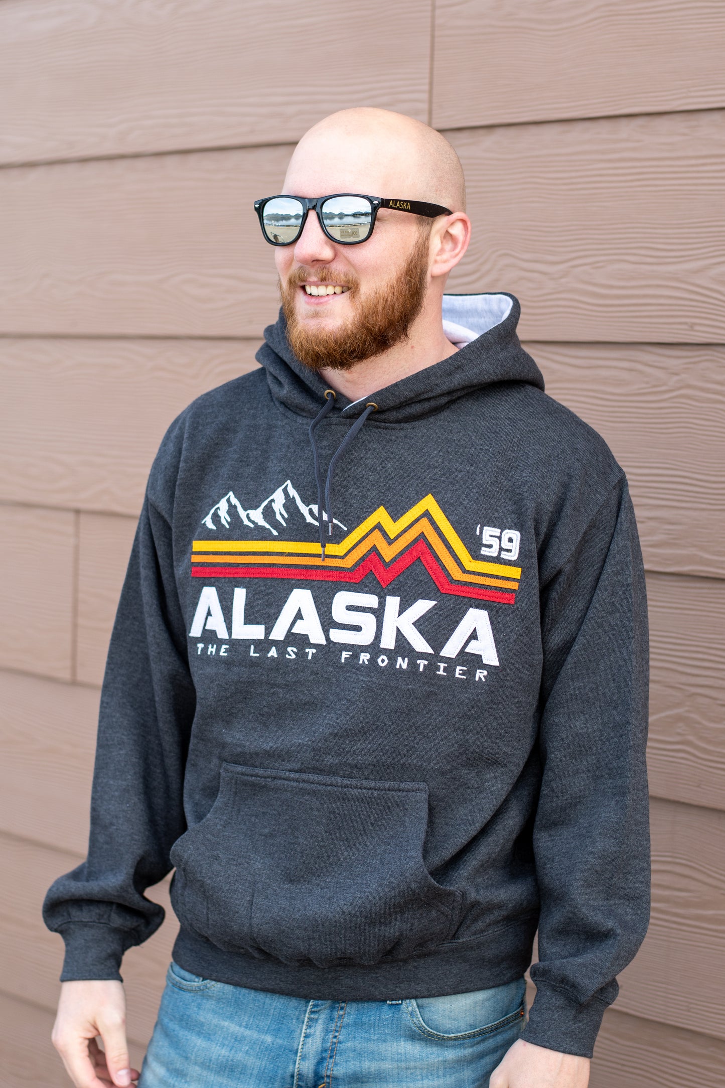Man wearing a grey hoodie with a red, yellow, and orange mountain outline with the text "Alaska, the last frontier"