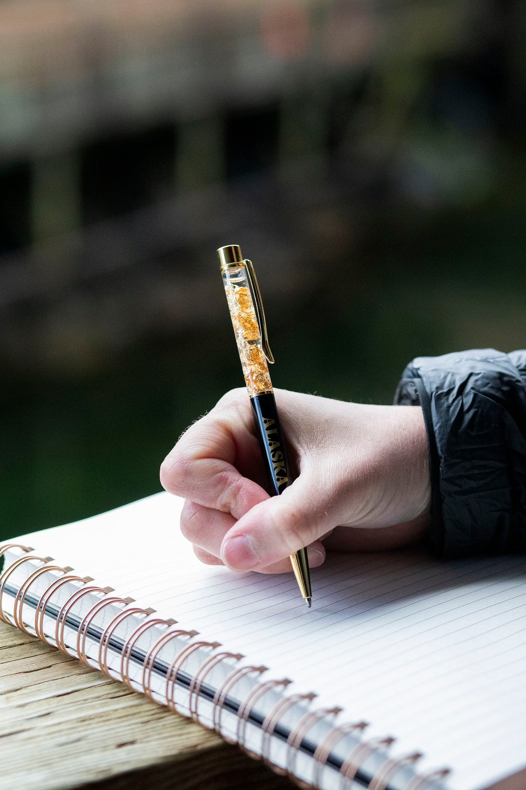 Hand holding a pen over a notebook with a blurred natural background