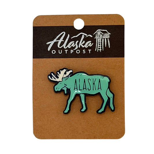 Green moose-shaped Alaska pin on a brown card with 'Alaska Outpost' branding.
