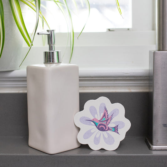 White soap dispenser on a bathroom counter with an eco-sponge featuring a colorful hummingbird design.