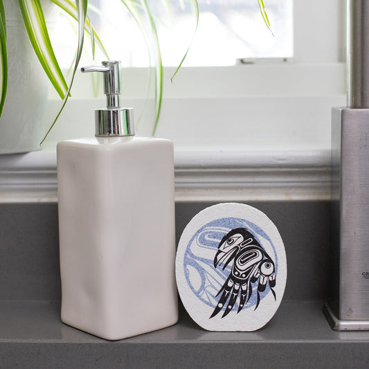White soap dispenser with an Eco-sponge with formline art of a raven and a moon, on a bathroom shelf.