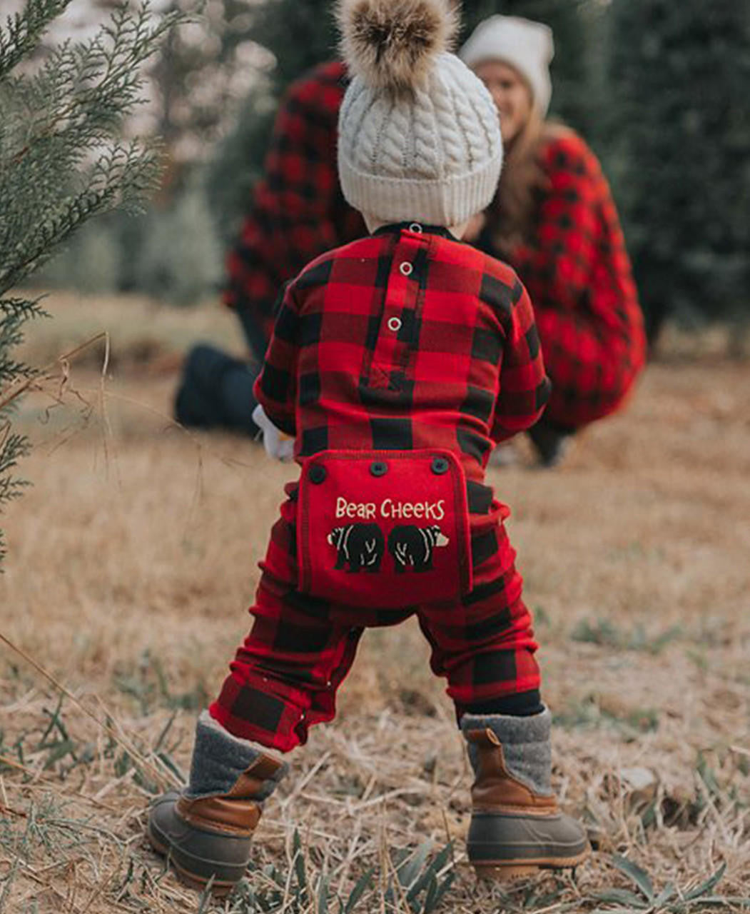Child wearing a red and black plaid outfit with 'Bear Cheeks' on the back in a tree farm setting.