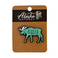 Green moose-shaped Alaska pin on a brown card with 'Alaska Outpost' branding.