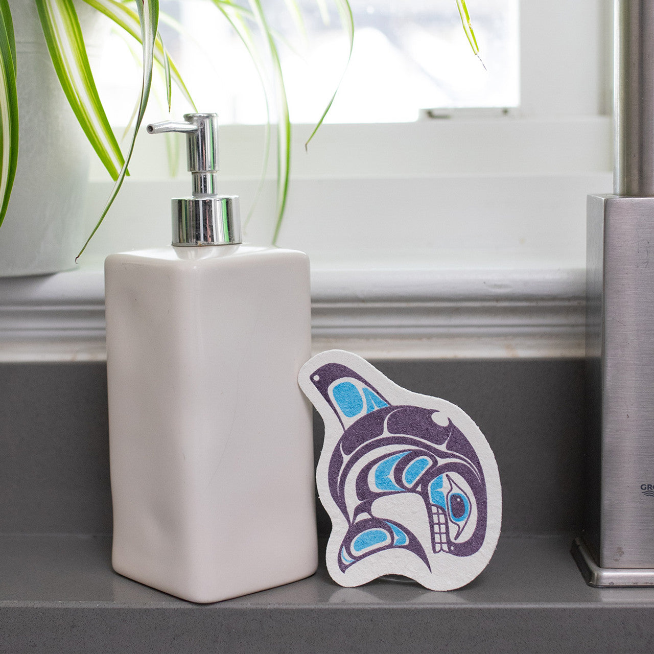 Soap dispenser on a bathroom counter with an eco sponge featuring a purple and blue form line whale design