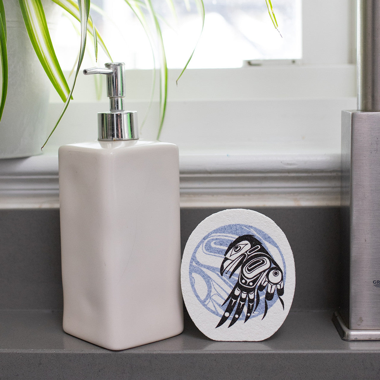 White soap dispenser with an Eco-sponge with formline art of a raven and a moon, on a bathroom shelf.