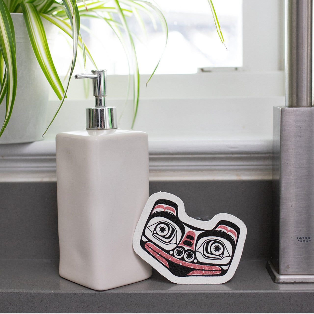 White soap dispenser with an Eco Sponge leaning on it, with a formline matriarch bear design 